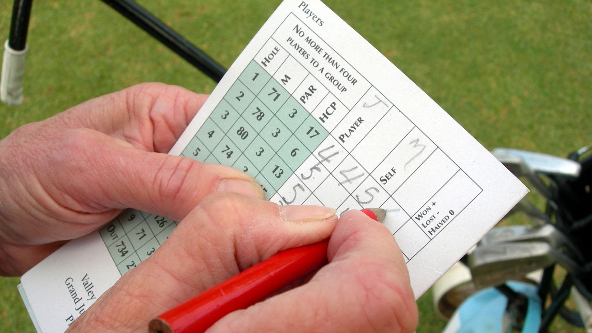 Close-up of hands holding a red pencil, expertly jotting down golf scoring terms on a scorecard amid the lush green.